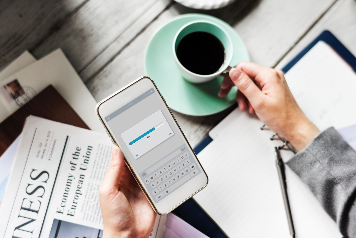 Hand holding smartphone showing a 72% progress bar while the other hand holds a cup of coffee over a notebook with a pen and a newspaper titled 'Economy of the European Union' nearby.