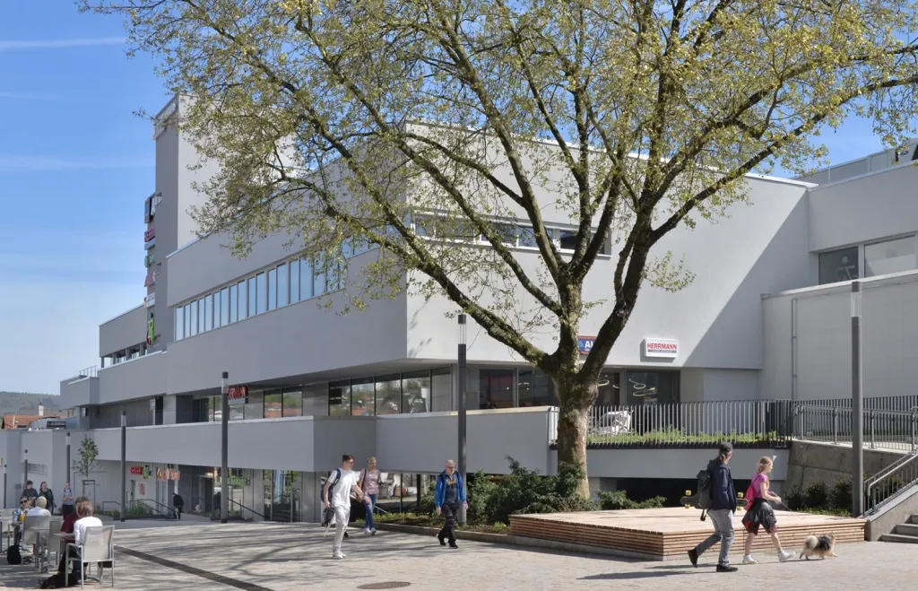 Modern multi-story building with large windows, a central tree, and people walking and sitting outside on a sunny day.